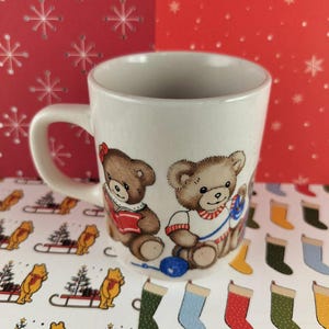 May include: White ceramic mug featuring two cartoon teddy bears, one reading a book and the other with a ball of yarn. The mug has a curved handle and is decorated with Christmas-themed patterns.