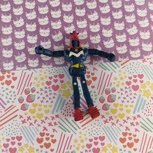 以下が含まれることがあります： 腕を広げた青と赤のロボットのおもちゃが、模様のある表面に立っています。ロボットは赤い頭、青い体、赤い足を持っています。背景には紫色の猫の模様と、ハート、星、イチゴを使ったカラフルなデザインがあります。