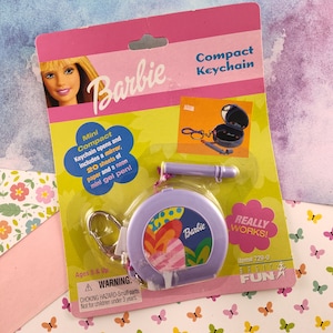 Puede incluir: Un llavero compacto Barbie morado con un espejo, 20 hojas de papel y un mini bolígrafo de gel de neón. El llavero tiene un diseño colorido con un corazón y la palabra "Barbie" en él. El embalaje dice "Really Works!" y "Basic Fun."