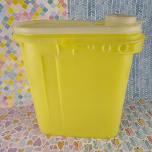 May include: A bright yellow plastic container with a matching lid. The container has a rectangular shape with rounded corners and a handle on the lid. The background features a colorful patterned design.