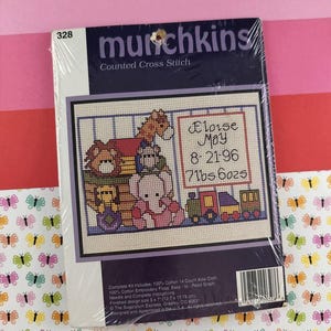May include: A sealed cross-stitch kit labeled "munchkins" with a finished design size of 5 x 7 inches. The design features a baby-themed scene with a train, elephant, and giraffe. The text includes the name "Eloise May" and birth details.