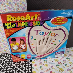 May include: RoseArt My First Impression 3-D Keepsake kit with a heart-shaped mold, paint, and a brush. The box shows a child's handprint in the mold with the name "Taylor" written above it.