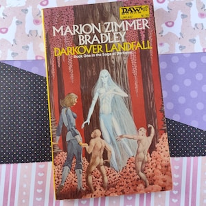 May include: A paperback book cover featuring a white background with a red, pink, and green illustration of a woman in a white dress, a man in a blue uniform, and a monkey-like creature. The book title is "Darkover Landfall" and the author is "Marion Zimmer Bradley".