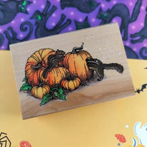 May include: Wooden stamp featuring a Halloween design with orange pumpkins and two squirrels. The stamp has a light wood grain. The background includes a purple pattern with black cat silhouettes.