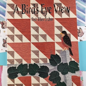 Op de afbeelding: Een boek getiteld "A Bird's Eye View" van Ruth Ellen Srubas. De omslag heeft een rood, wit en bruin patchwork-ontwerp met een vogel die op een tak zit met groene bladeren.