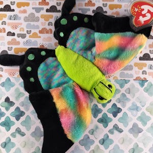 May include: A soft butterfly toy with black, green, and rainbow wings. The butterfly has a green body and black antennae. The wings are a mix of pink, yellow, blue, and green. A red tag is attached.