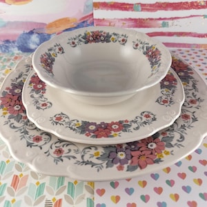 May include: A set of three white ceramic plates with a floral design in shades of pink, purple, and gray. The plates have a scalloped edge and are decorated with a delicate floral pattern.