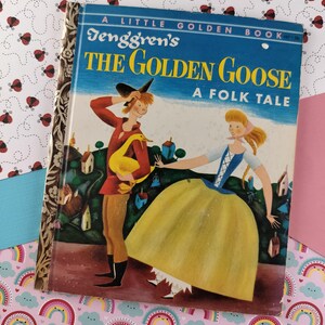 Pode incluir: Um pequeno livro dourado intitulado "A Galinha dos Ovos de Ouro" de Tenggren's. A capa apresenta uma ilustração colorida de um homem e uma mulher com uma gansa dourada.