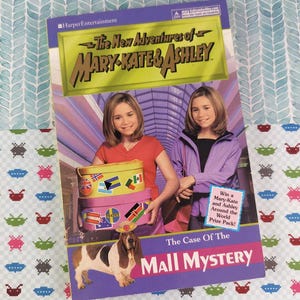 Puede incluir: Portada del libro "Las nuevas aventuras de Mary-Kate & Ashley: El caso del misterio del centro comercial". La portada presenta a las gemelas Olsen, maletas apiladas con pegatinas de banderas y un perro basset. El libro es morado, rosa y amarillo.