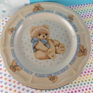 May include: A beige ceramic plate featuring a teddy bear design. The plate has a blue border with small teddy bear illustrations and a central image of a teddy bear with a blue bow. The plate has a scalloped edge.