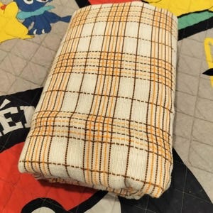 May include: Folded white and tan plaid blanket. The blanket has a repeating pattern of thin brown and orange lines. The blanket is folded and resting on a patterned quilt with cartoon characters.