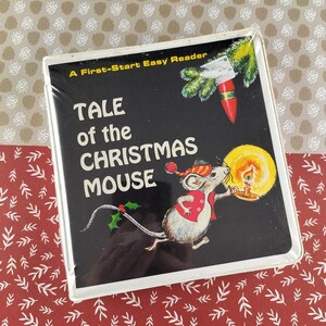 Könnte beinhalten: Ein Kinderbuch mit dem Titel "Tale of the Christmas Mouse" mit schwarzem Einband. Das Cover zeigt eine Cartoon-Maus, die eine Kerze, einen Weihnachtsbaumzweig und eine rot-weiße Verzierung hält. Das Buch befindet sich in einer transparenten Kunststoffhülle.