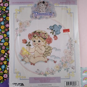 Puede incluir: Un kit de punto de cruz contado con un querubín con una corona de flores y alas, sentado en una nube con un pájaro azul. El kit incluye tela Aida de 18 hilos, hilo de algodón, cuentas, una aguja, un gráfico e instrucciones. El tamaño terminado es de 17,8 cm por 17,8 cm, y el tamaño del marco es de 48,3 cm por 22,9 cm. El kit se titula "Rainbow Dreams" y forma parte de la colección Dreamsicles.