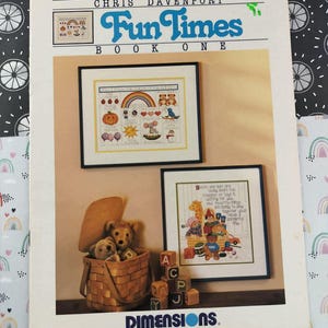 May include: A vintage "Fun Times Book One" cross-stitch pattern book by Chris Davenport. The cover features framed cross-stitch samplers with colorful designs, including a rainbow, teddy bears, and a basket of toys. A Dimensions logo is visible.
