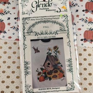 Puede incluir: Un patrón de pintura termoadhesiva Glenda Fashion Patterns con un diseño otoñal. El diseño incluye una casa para pájaros, girasoles, calabazas y el texto "Autumn Home". El paquete está sellado en plástico.
