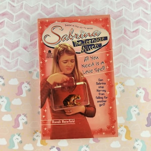 Op de afbeelding: Een paperback boek getiteld "Sabrina The Teenage Witch: All You Need Is a Love Spell". De rode cover heeft witte tekst en een foto van een jonge vrouw die een kikker in een doorzichtige bak vasthoudt.