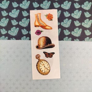 Peut inclure: Une planche d'autocollants avec des illustrations vintage. Elle comprend une botte beige, un chapeau melon marron, une montre de poche dorée, un papillon et deux fleurs. Les illustrations ont une teinte sépia.