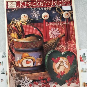 May include: A decorative winter-themed artwork featuring a watering can, heart-shaped box, and snowman-topped sticks. The artwork includes the text "Kracker Jack Krittters" and is adorned with snowflakes and red berries. The color palette includes red, blue, and green.
