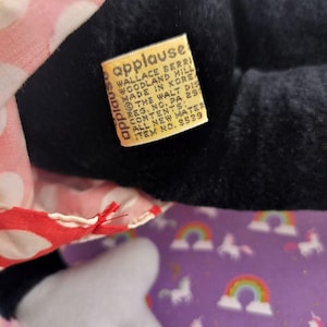 May include: Close-up of a black plush toy with a red and white polka dot dress. A yellow tag reads "applause" and includes the manufacturer's information. The toy is on a purple background with unicorns and rainbows.