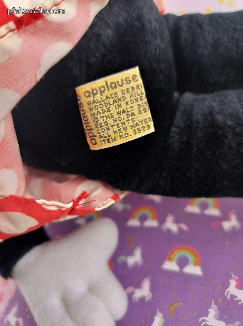 May include: Close-up of a black plush toy with a red and white polka dot dress. A yellow tag reads "applause" and includes the manufacturer's information. The toy is on a purple background with unicorns and rainbows.