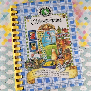 Puede incluir: Un libro de cocina encuadernado en espiral titulado "Celebrate Spring" con una cubierta de cuadros azules y encuadernación en espiral amarilla. La portada presenta una colorida ilustración de una escena primaveral con un gato, flores y limones. El logotipo de Gooseberry Patch es visible.
