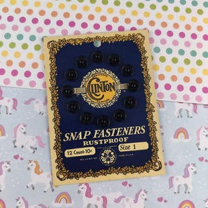 Puede incluir: Una tarjeta de broches de presión negros, talla 1, con el texto "SNAP FASTENERS RUSTPROOF" y "12 Count-10". La tarjeta tiene un fondo azul marino con detalles dorados y la marca "CLINTON".