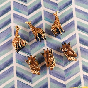 Könnte beinhalten: Eine Sammlung von Giraffenfiguren und Giraffenkopf-förmigen Anstecknadeln. Die Figuren sind braun und beige mit detaillierten Flecken, während die Anstecknadeln ein ähnliches Farbschema aufweisen. Die Artikel sind auf einem blau-weißen Zickzackmuster angeordnet.
