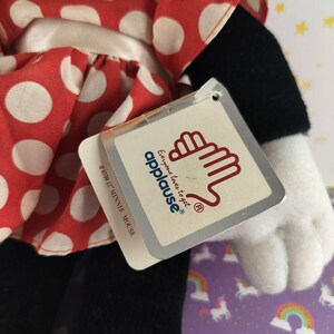 May include: A Minnie Mouse plush doll with a red and white polka dot dress. The doll has black pants, a white glove, and a tag that reads "Everyone loves to get applause&reg;". The tag also says "#8539 17" MINNIE MOUSE."