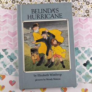 Pode incluir: Um livro azul intitulado "O furacão de Belinda" de Elizabeth Winthrop com ilustrações de Wendy Watson. A capa apresenta uma ilustração colorida de três pessoas caminhando na praia com um cão.