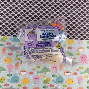 Puede incluir: Un juguete Happy Meal sellado de McDonald's, con un paquete morado que dice "HAPPY BIRTHDAY Happy Meal" y "Cabbage Patch Kids". El fondo es un patrón azul claro con criaturas marinas.