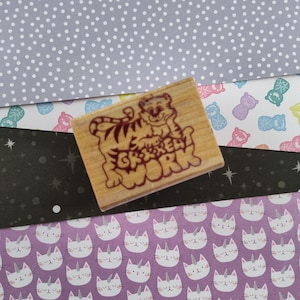 May include: A wooden stamp with a cartoon tiger and the text "Get to Work" in red lettering. The stamp is on top of patterned paper with white polka dots, pastel colored flowers, and white cats with silver horns.