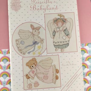 Peut inclure: Un livre de patrons de point de croix intitulé "Priscilla's Babyland" avec des motifs de Gloria & Pat. La couverture présente trois images de point de croix d'un lapin, d'un ange et d'un ours en peluche, tous avec des ailes d'ange. Le livre est sur fond rose.