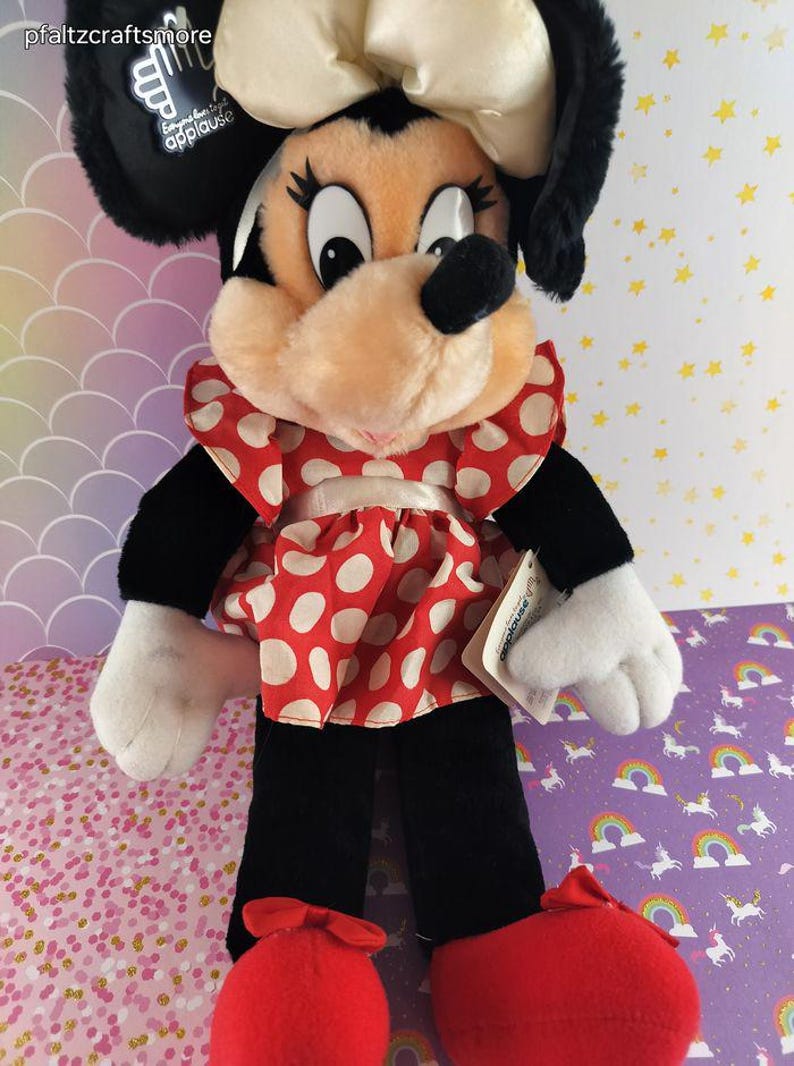 May include: A plush Minnie Mouse doll with a cream-colored bow, black ears, and a red and white polka-dot dress. The doll has black pants, white gloves, and red shoes. The tag reads "applause".