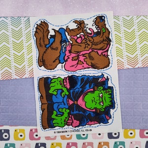 May include: Two vintage stickers featuring cartoon illustrations of a werewolf and Frankenstein's monster. The werewolf is brown with a pink shirt and blue pants. The monster is green with a blue shirt and pink pants. Both stickers have a blue dotted outline and the text "1984 MARK 1 CHICAGO, ILL ISS-39".