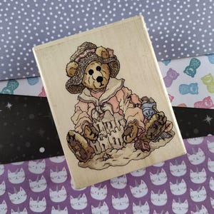 May include: A wooden rubber stamp featuring a cartoon bear wearing a pink dress and a hat, sitting on a beach with a sandcastle and seashells.