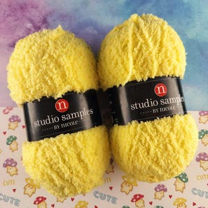May include: Two fluffy yellow yarn skeins with black labels that read "studio samples by Nicole." The yarn is on a patterned surface with mushroom illustrations and the word "CUTE." The yarn is a bright, sunny yellow.