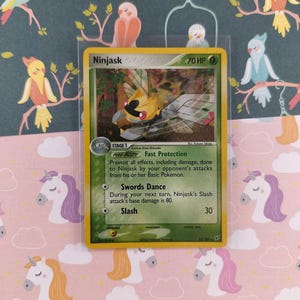 Pode incluir: Uma carta colecionável Pokémon Ninjask em uma manga protetora. A carta apresenta uma criatura insetóide amarela e preta com asas. O fundo é um padrão colorido de pássaros e unicórnios.