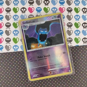 May include: A Golbat lv.22 Pokémon card in a protective sleeve. The card features a blue bat-like creature with open wings and the text "Pulse Search". The background has a pattern of skulls and hearts.