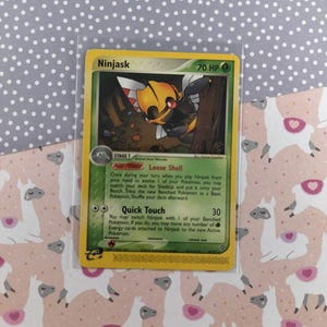 Pode incluir: Um cartão Pokémon Ninjask amarelo e vermelho do Jogo de Cartas Colecionáveis Pokémon. O cartão apresenta uma ilustração de Ninjask, um Pokémon do tipo inseto, com o texto "Ninjask" e "70 PV" no topo do cartão. O cartão também apresenta o texto "Casca Solta" e "Toque Rápido" com suas descrições de ataque correspondentes.
