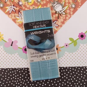 Könnte beinhalten: Eine Packung blaues, weiches und leicht zu verarbeitendes Saumband von Wrights. Die Verpackung ist schwarz mit weißem Text und trägt die Aufschrift "Soft and Easy Hem Tape" und "Wrights". Außerdem steht "39c" und "3 yds" darauf. Die Verpackung enthält ein blaues Saumband-Muster.