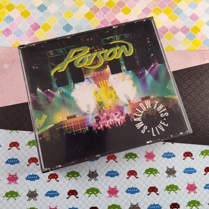 May include: A black CD case with a silver band around the edge. The front of the case features a black and white image of a concert stage with the band Poison's name in white letters. The text "Swallow This Live" is printed in white letters on a black circle.