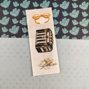 Peut inclure: Marque-page avec des illustrations de lunettes à monture dorée, d'une malle noire et dorée et d'une pile de papiers avec une plume. Le fond présente un motif de formes de papillons bleu-vert sur fond bleu foncé.