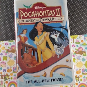 Puede incluir: Una cinta VHS de Pocahontas II: Viaje a un nuevo mundo de Disney. La portada presenta a Pocahontas con un vestido amarillo, un mapache y otros personajes en un barco. El título está en rojo y dorado, con la frase "THE ALL-NEW MOVIE!"