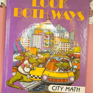 May include: Purple book cover with the title "LOOK BOTH WAYS CITY MATH". The cover features a cartoon illustration of a city scene with a yellow taxi cab and several alligators.
