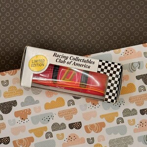May include: A red toy car in a clear plastic box with a black and white checkered pattern on the side. The box has a white label with the text "Racing Collectables Club of America" and "Limited Edition".