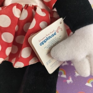 May include: A plush doll featuring a red dress with white polka dots. The doll has black legs and arms, a white glove, and a tag that reads "Everyone loves to get applause". The doll is a collectible item.