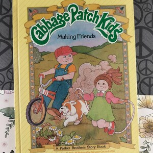 Op de afbeelding: Een vintage "Cabbage Patch Kids" verhalenboek getiteld "Making Friends". De cover bevat illustraties van kinderen, een hond en een fiets. Het boek is een Parker Brothers Story Book, geschreven door Kathleen N. Daly en geïllustreerd door Maryann Cocca-Leffler.