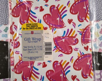 Pode incluir: Um pacote de duas folhas de papel de embrulho branco com um padrão de corações rosa. Os corações são delineados em cores do arco-íris. O pacote inclui o texto "package includes two sheets for the price of one" e "American Greetings Gift Wrap".