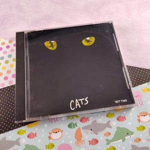 May include: A black CD case with a black cat's face and yellow eyes. The text "CATS" is printed in white on the case. The text "ACT TWO" is printed in white in the top right corner of the case.