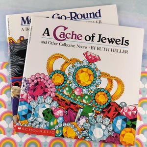 Puede incluir: Dos libros infantiles apilados. El libro superior se titula "A Cache of Jewels" de Ruth Heller, con una ilustración de portada de una corona enjoyada y piedras preciosas. El libro inferior se titula "Go-Round".
